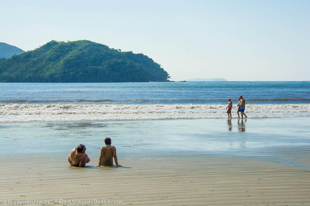 Imagem de um casal sentado na beira da praia e amigos caminhando.